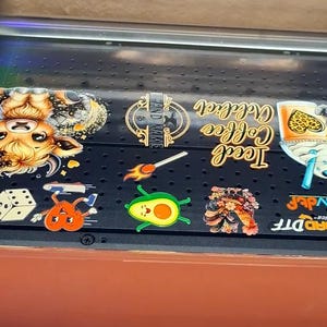 May include: A close-up of a black surface with various colourful stickers. The stickers feature a cartoon cow, a matchstick, an avocado, a flower, a dice, a heart, and a glass of iced coffee. The text "Iced Coffee" and "Brand Name" are visible on the stickers.