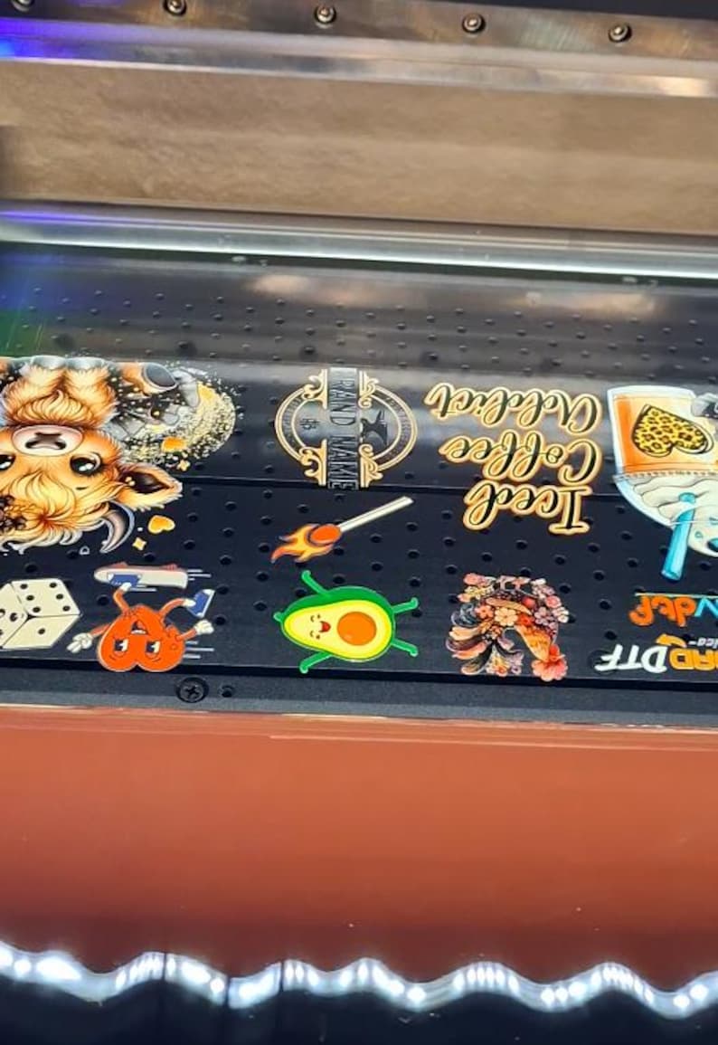 May include: A close-up of a black surface with various colourful stickers. The stickers feature a cartoon cow, a matchstick, an avocado, a flower, a dice, a heart, and a glass of iced coffee. The text "Iced Coffee" and "Brand Name" are visible on the stickers.