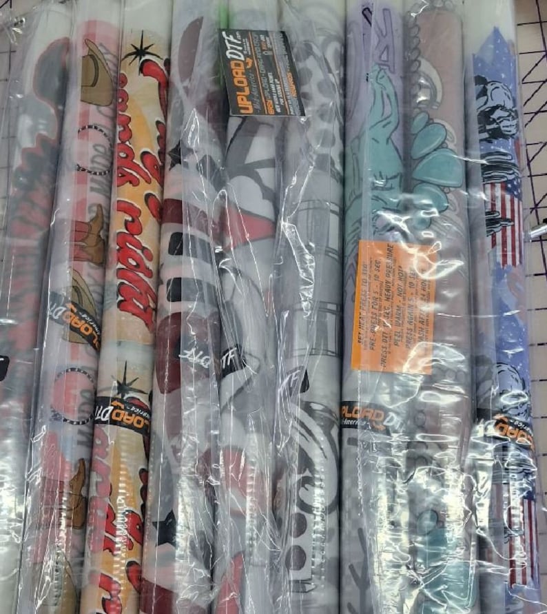 Pode incluir: Um close-up de v&aacute;rios rolos de folhas de decalque de vinil, embrulhados em pl&aacute;stico transparente. Os decalques apresentam v&aacute;rios desenhos, incluindo uma bota de cowboy, um carro antigo e um tema patri&oacute;tico. O texto "UPLOAD DTF" &eacute; vis&iacute;vel em um dos rolos.