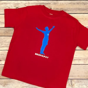 May include: Red t-shirt with a blue silhouette of a soccer player with the number 15 on the jersey and the text #EQUALPAY.