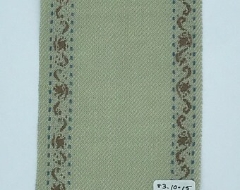 May include: A light green fabric swatch with a brown and blue patterned border. The fabric has a woven texture. A price tag is attached to the bottom right corner of the swatch. The price tag reads "$3.10/5 $22."