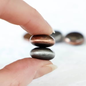 May include: Two metallic, oval-shaped beads are held between fingers. The top bead has a copper hue, while the bottom one is silver. Other beads are slightly out of focus in the background. The beads appear to be smooth and polished.