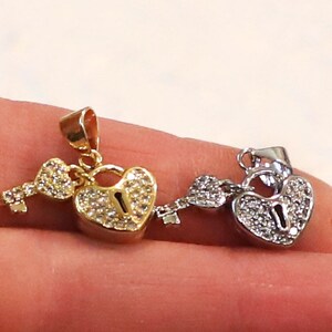 Puede incluir: Dos colgantes de coraz&oacute;n con candado y llave, uno dorado y otro plateado, con detalles de cristal transparente.