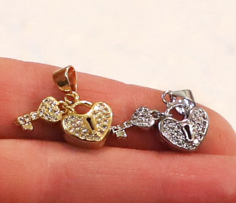 Puede incluir: Dos colgantes de coraz&oacute;n con candado y llave, uno dorado y otro plateado, con detalles de cristal transparente.
