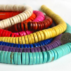 May include: A close-up of a multi-colored strand of round, flat beads. The beads are arranged in a rainbow pattern, starting with white, then orange, red, pink, brown, yellow, blue, purple, and turquoise. The beads are made of natural stone and have a slightly rough texture.