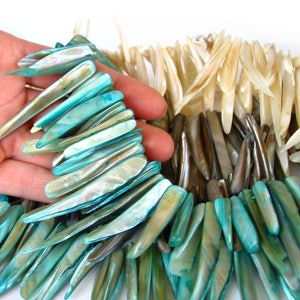 Puede incluir: Un collar hecho a mano con piezas de concha iridiscentes de color turquesa, marrón y crema. Las conchas son alargadas y están dispuestas en un diseño en capas. El collar es sostenido por una mano, mostrando la textura y las variaciones de color.