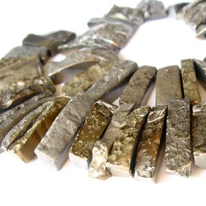 Op de afbeelding: Een close-up van een ketting gemaakt van lange, rechthoekige, metalen kralen. De kralen zijn een mix van zilveren en gouden tinten, met een ruw, getextureerd oppervlak.