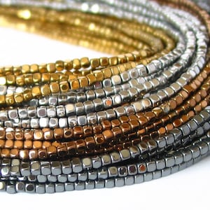 May include: Close-up of multiple strands of square beads in various metallic colors, including gold, silver, bronze, and gunmetal gray. The beads are small and uniform, creating a textured, layered effect. Ideal for jewelry making.