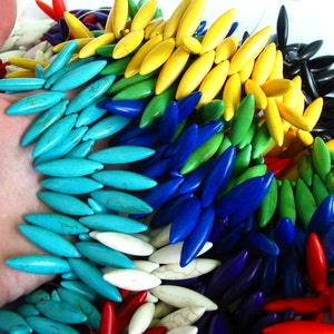 Puede incluir: Un collar multicolor compuesto por cuentas alargadas de forma ovalada. Las cuentas están dispuestas en un patrón circular, mostrando un espectro de colores que incluye turquesa, amarillo, verde, azul y rojo. El collar se muestra sobre un fondo blanco.