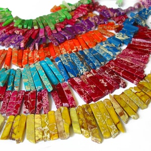 Könnte beinhalten: Eine bunte Auswahl an langen, rechteckigen, polierten Edelsteinperlen. Die Perlen sind fächerförmig angeordnet, wobei die Farben von Grün über Rot nach Gelb übergehen. Die Perlen bestehen aus verschiedenen Materialien, darunter Türkis, Jaspis und Achat.