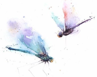 May include: A watercolor painting featuring two dragonflies in flight, rendered with splashes of blue, purple, and orange pigment. The insects have delicate, elongated bodies and translucent wings. The word Sweet is written in cursive in the bottom right corner.