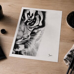 May include: A black and white charcoal drawing on paper depicting the close-up face of a tiger with detailed stripes and whiskers. The artwork features a signature in the bottom right corner and is surrounded by art supplies including black paint.