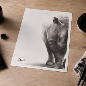 May include: A black and white charcoal drawing on paper depicting a rhinoceros, positioned on the right side of the frame. The artwork features a signature in the bottom left corner and is surrounded by art supplies including paint and brushes.