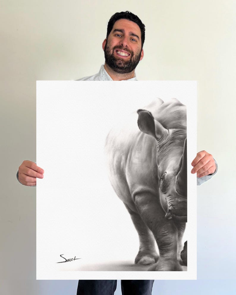 May include: A rectangular art print featuring a detailed black and white charcoal-style drawing of a rhinoceros. The animal is positioned on the right side of the white paper, with a signature in the bottom left corner that reads Sweet.