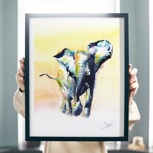 May include: A watercolor painting of an elephant in a yellow, orange, and blue color scheme. The elephant is walking towards the viewer with its trunk raised. The painting is framed in a black frame.