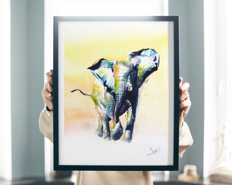 May include: A watercolor painting of an elephant in a yellow, orange, and blue color scheme. The elephant is walking towards the viewer with its trunk raised. The painting is framed in a black frame.
