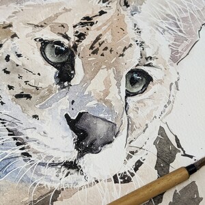 Op de afbeelding: Close-up aquarel schilderij van het gezicht van een serval. De serval heeft grote, groene ogen en een zwarte neus. Het schilderij is gemaakt in tinten bruin, grijs en wit.