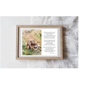 May include: A framed print featuring a photograph of two young girls lying in grass. The print includes a poem with the text "Girls, you were born to run..." in a wooden frame. The background is a white surface.