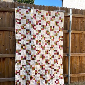 May include: A patchwork quilt with a white background and squares of pink, green, and gold fabric. The quilt is hanging on a wooden fence.