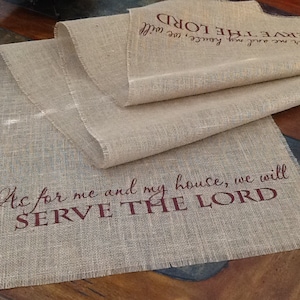 May include: A burlap table runner with the text "As for me and my house, we will SERVE THE LORD" printed in brown. The runner is folded in half and has frayed edges.