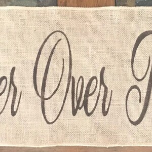 Puede incluir: Una pancarta rectangular de arpillera color canela con el texto marrón oscuro "His Banner Over Me Is Love" escrito en una fuente cursiva. La tela tiene una textura tejida visible y los bordes son rectos, mostrando un color marrón claro.