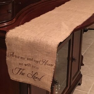 May include: A burlap table runner with the text "As for me and my house we will serve The Lord" printed in brown. The runner is on a wooden table with a glass door.