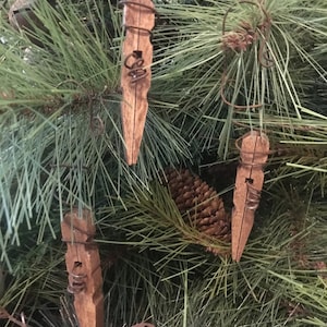 以下が含まれることがあります： 緑の松の枝に、ワイヤーハンガーが付いた素朴な木製の洗濯ばさみオーナメントが3つぶら下がっています。