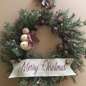 Può includere: Una ghirlanda natalizia festosa con rami di sempreverdi finti, piccole campane e bacche rosse. Una figura di pupazzo di neve con una sciarpa a quadri è presente a sinistra. Uno striscione di iuta in basso recita "Merry Christmas". Un fiocco a quadri è in alto.