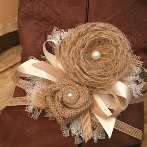 Pode incluir: Uma bota de couro marrom com um enfeite de flor de juta e renda. A flor tem uma pérola branca no centro e está amarrada com uma fita de cetim branco.