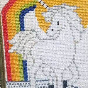 May include: A framed cross-stitch of a white unicorn standing in front of a rainbow. The unicorn has a gold horn and a gray mane and tail.
