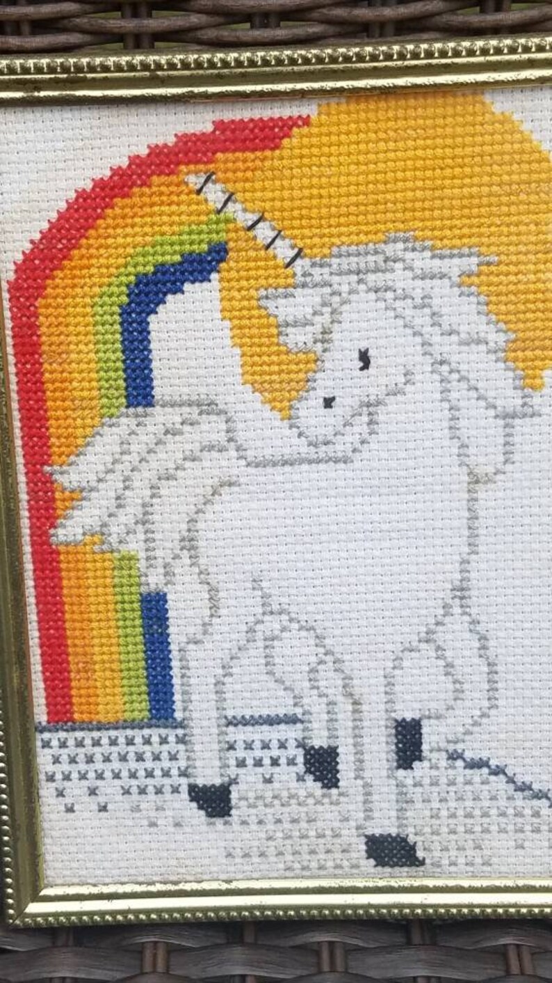 May include: A framed cross-stitch of a white unicorn standing in front of a rainbow. The unicorn has a gold horn and a gray mane and tail.