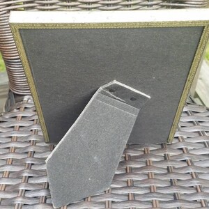 May include: A gold-colored picture frame with a black mat and a black cardboard easel stand. The frame is sitting on a woven wicker surface.