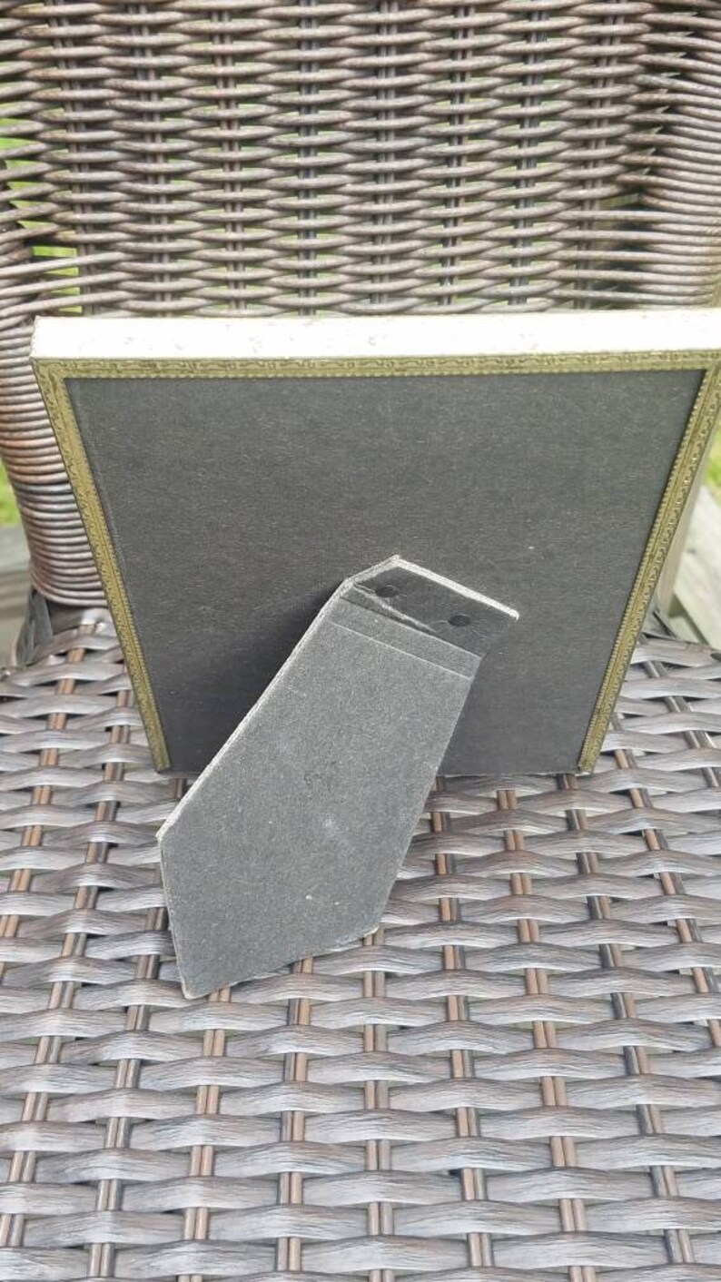 May include: A gold-colored picture frame with a black mat and a black cardboard easel stand. The frame is sitting on a woven wicker surface.