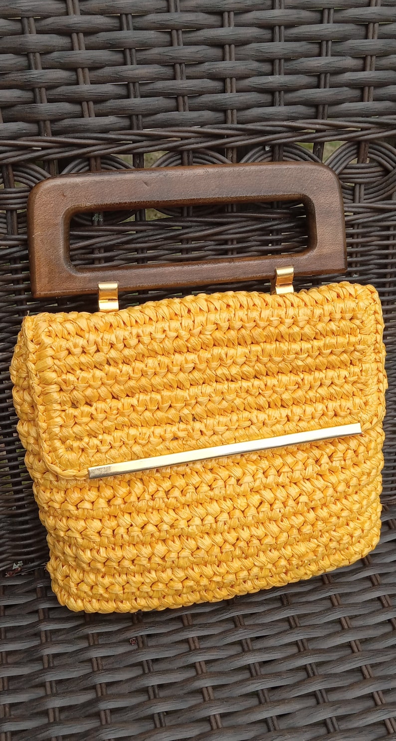Pode incluir: Uma bolsa de m&atilde;o de croch&ecirc; amarela com uma al&ccedil;a de madeira e uma barra de metal dourado na frente.