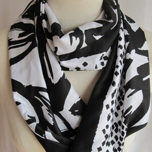 May include: A black and white infinity scarf with a bold abstract design. The scarf features a repeating pattern of black brushstrokes and geometric shapes against a white background. The scarf is draped around a mannequin's neck.