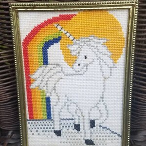 May include: A framed cross-stitch of a white unicorn standing in front of a rainbow. The unicorn has a gold horn and is standing on a white cloud.