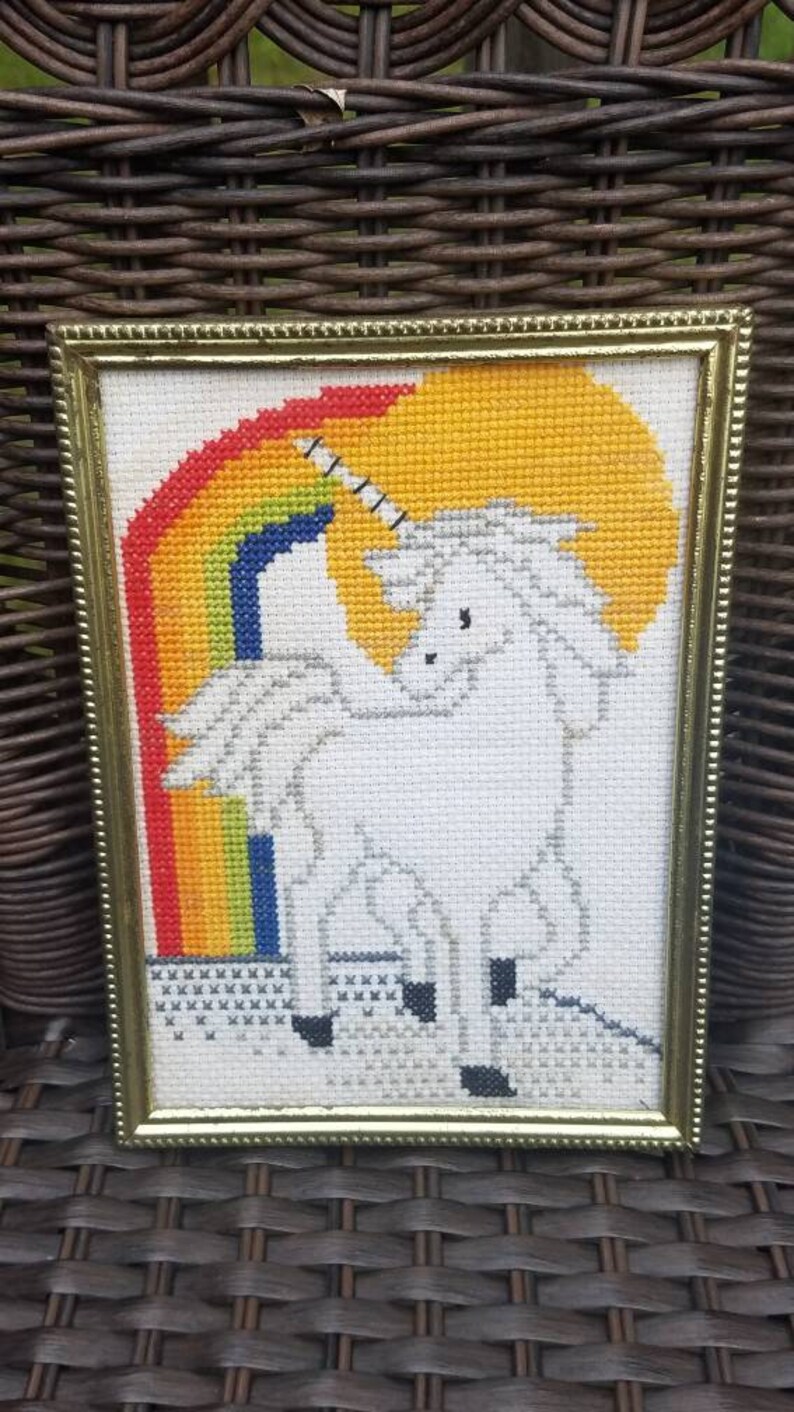 May include: A framed cross-stitch of a white unicorn standing in front of a rainbow. The unicorn has a gold horn and is standing on a white cloud.