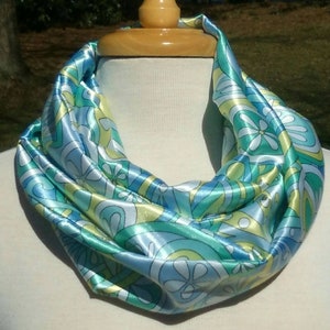 May include: A silky infinity scarf in shades of blue, green, and yellow, with a floral and abstract pattern. The scarf is draped around a mannequin neck, showcasing its flowing design and vibrant colors. The scarf is made of a shiny, smooth fabric.