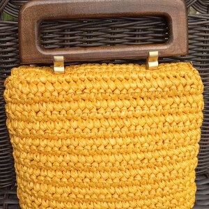 Pode incluir: Uma bolsa de m&atilde;o de croch&ecirc; amarela com al&ccedil;a de madeira. A bolsa tem uma textura tecida e fecha com um fecho dourado.