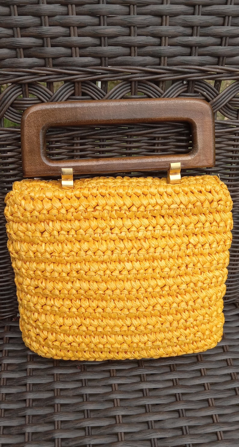 Pode incluir: Uma bolsa de m&atilde;o de croch&ecirc; amarela com al&ccedil;a de madeira. A bolsa tem uma textura tecida e fecha com um fecho dourado.