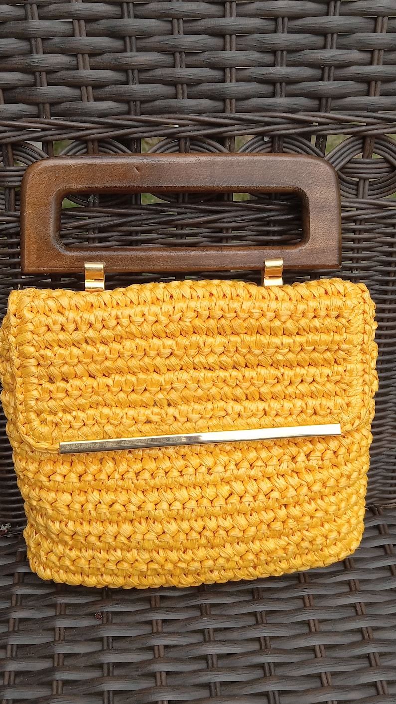 Pode incluir: Uma bolsa de m&atilde;o de croch&ecirc; amarela com uma al&ccedil;a de madeira e um detalhe em metal dourado.