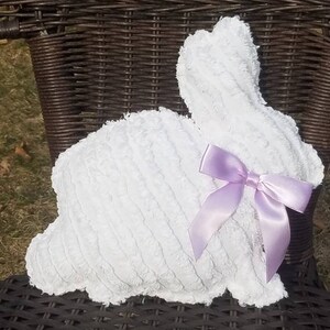 May include: A white plush bunny-shaped pillow with a lavender satin bow. The pillow has a textured surface and is sitting on a dark brown wicker chair. The bunny pillow is a decorative item.