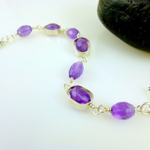 K&ouml;nnte beinhalten: Ein silbernes Armband mit einem Schiebeverschluss zeigt facettierte Amethyst-Edelsteine. Das Armband liegt auf einer wei&szlig;en Oberfl&auml;che.