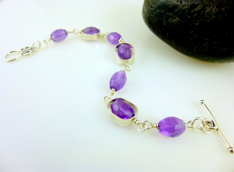 K&ouml;nnte beinhalten: Ein silbernes Armband mit einem Schiebeverschluss zeigt facettierte Amethyst-Edelsteine. Das Armband liegt auf einer wei&szlig;en Oberfl&auml;che.