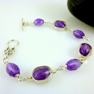 K&ouml;nnte beinhalten: Ein silbernes Kettenarmband mit einem Schiebeverschluss zeigt facettierte Amethyst-Edelsteine in ovalen und quadratischen Formen.