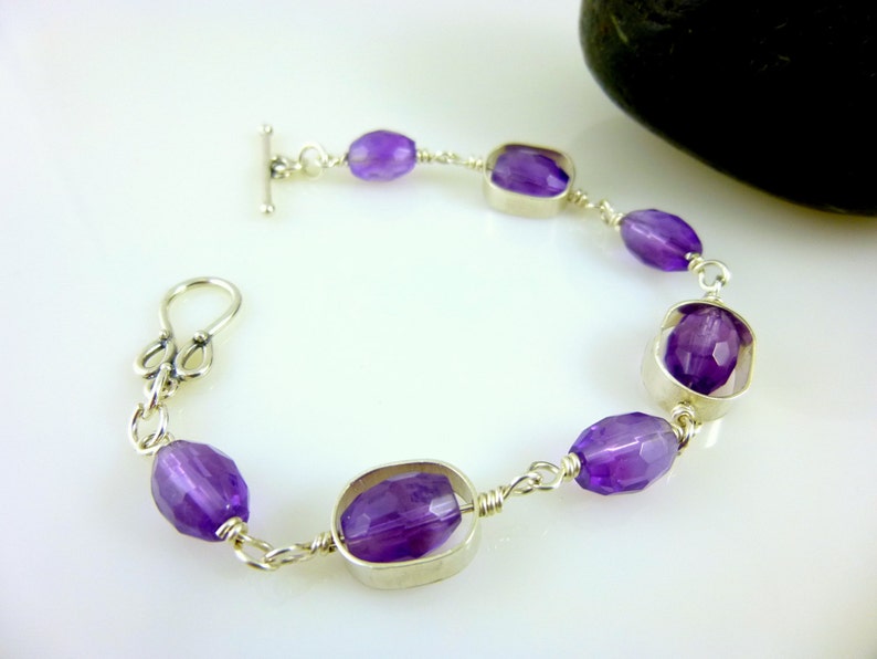 K&ouml;nnte beinhalten: Ein silbernes Kettenarmband mit einem Schiebeverschluss zeigt facettierte Amethyst-Edelsteine in ovalen und quadratischen Formen.