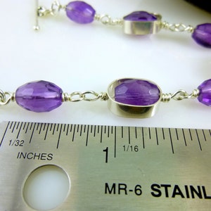 K&ouml;nnte beinhalten: Ein silbernes Kettenarmband mit violetten Amethyst-Edelsteinen. Das Armband wird an einem Lineal gemessen und zeigt, dass es etwas mehr als 2,5 cm lang ist.