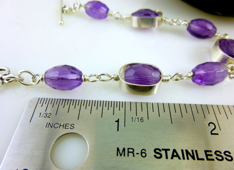 K&ouml;nnte beinhalten: Ein silbernes Kettenarmband mit violetten Amethyst-Edelsteinen. Das Armband wird an einem Lineal gemessen und zeigt, dass es etwas mehr als 2,5 cm lang ist.