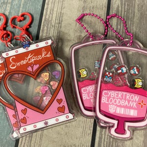 May include: Two pink plastic keychains. One keychain is shaped like a heart and has the text "Sweetsparks" on it. The other keychain is shaped like a blood bag and has the text "Cybertron Bloodbank" on it.