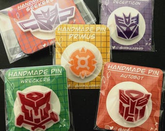 May include: Five different Transformers enamel pins, each with a different character and color scheme. The pins are packaged in clear plastic bags with a white background and colorful text that reads "Handmade Pin" and the name of the character. The characters are Seekers, Decepticon, Optimus Prime, Wreckers, and Autobot.